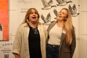 Two women laugh together in front of a wall decorated with flying Canada geese illustrations and Gander branding, appearing candid and joyful.