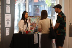 A woman stands behind a small table speaking to two attendees at an event. The table has a laptop, pens, and flyers promoting Gander’s Creator Program, with posters on the wall beside her. Bright light from the gallery window illuminates the scene.