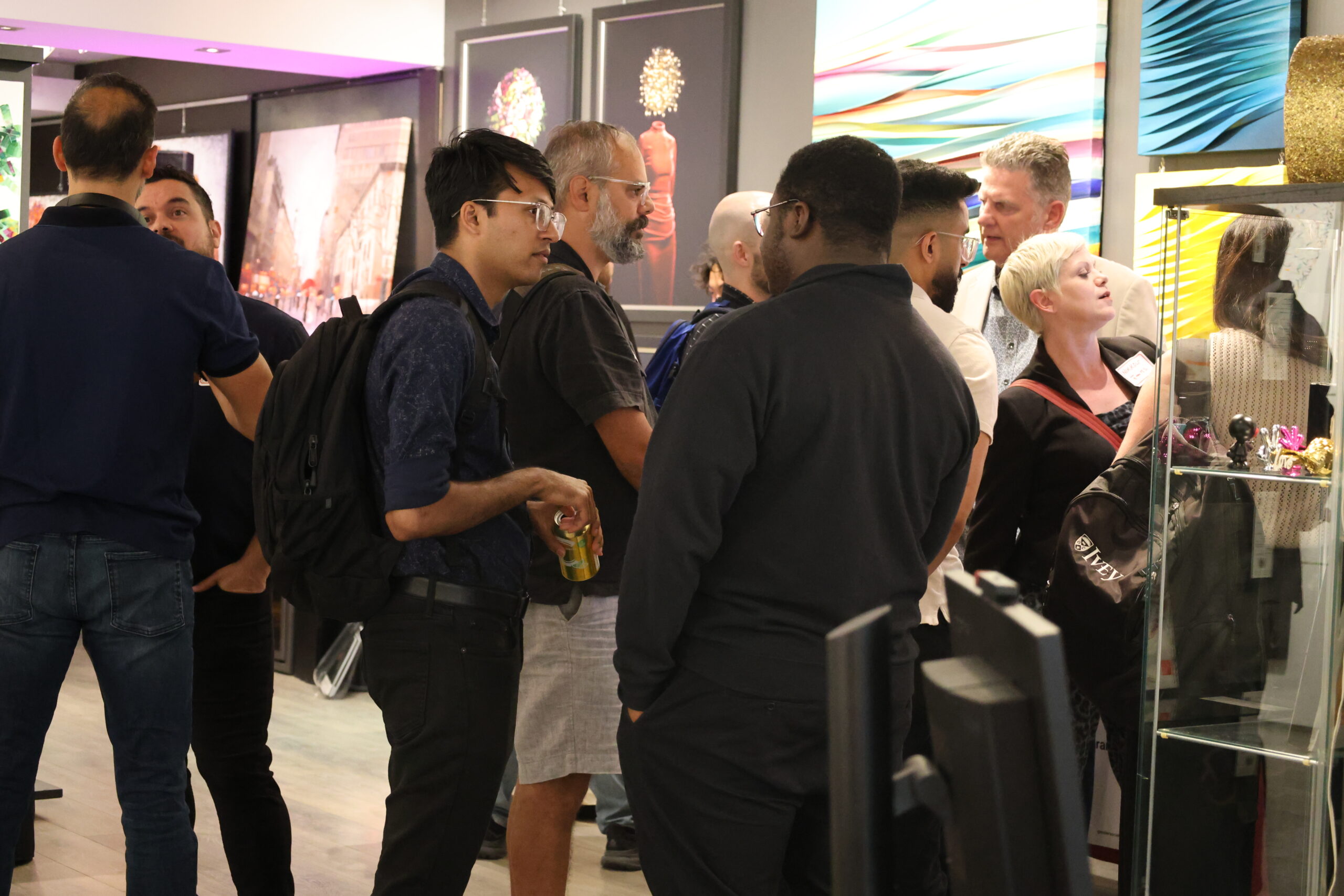 A group of people gather and talk inside an art gallery, standing near colourful abstract paintings and a glass display case. The atmosphere is lively and conversational.