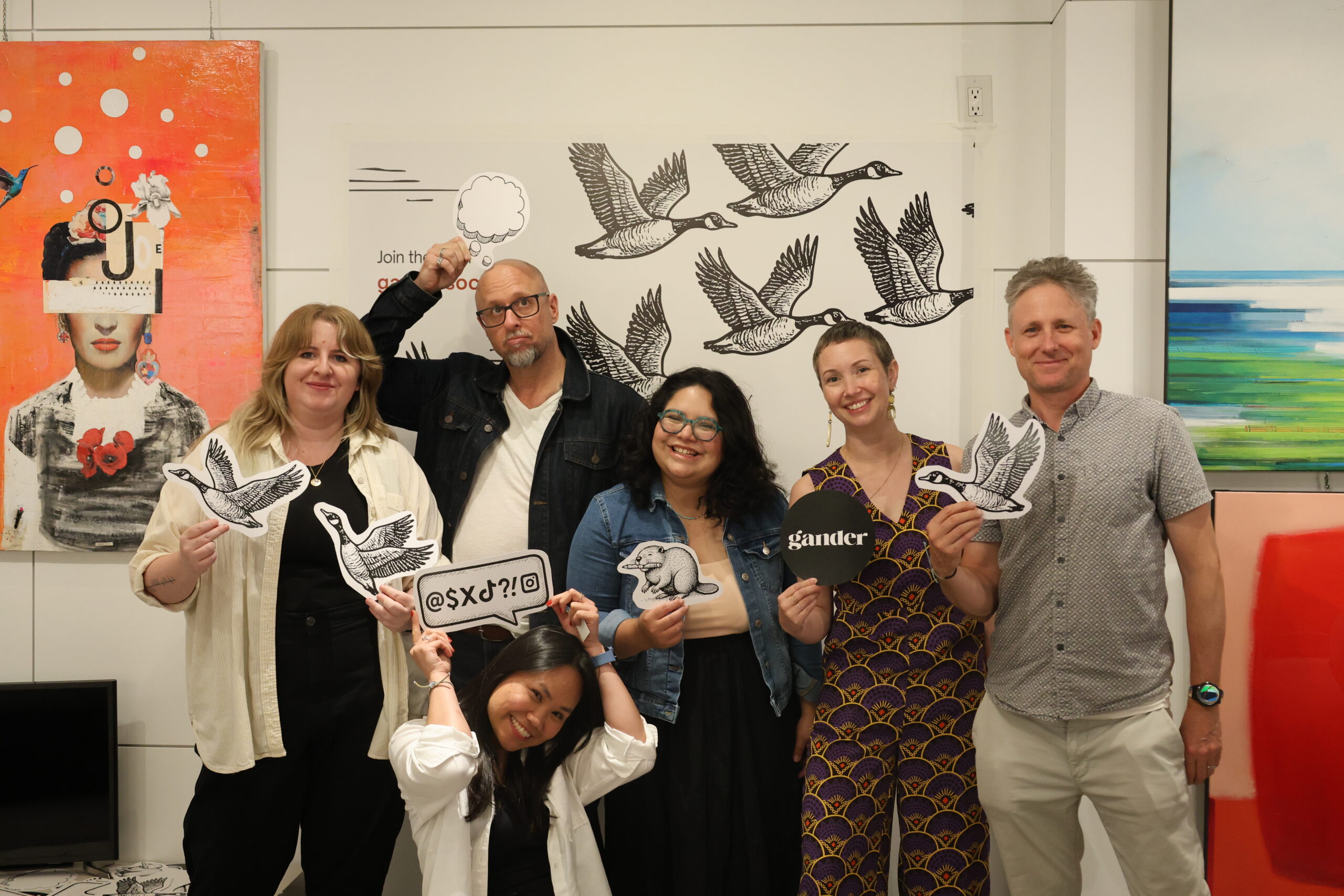 Six people stand together in front of artwork and a wall decorated with flying Canada geese illustrations. They are smiling and holding up playful cut-out props, including geese, a beaver, a speech bubble, the word “gander,” and a sign with symbols.