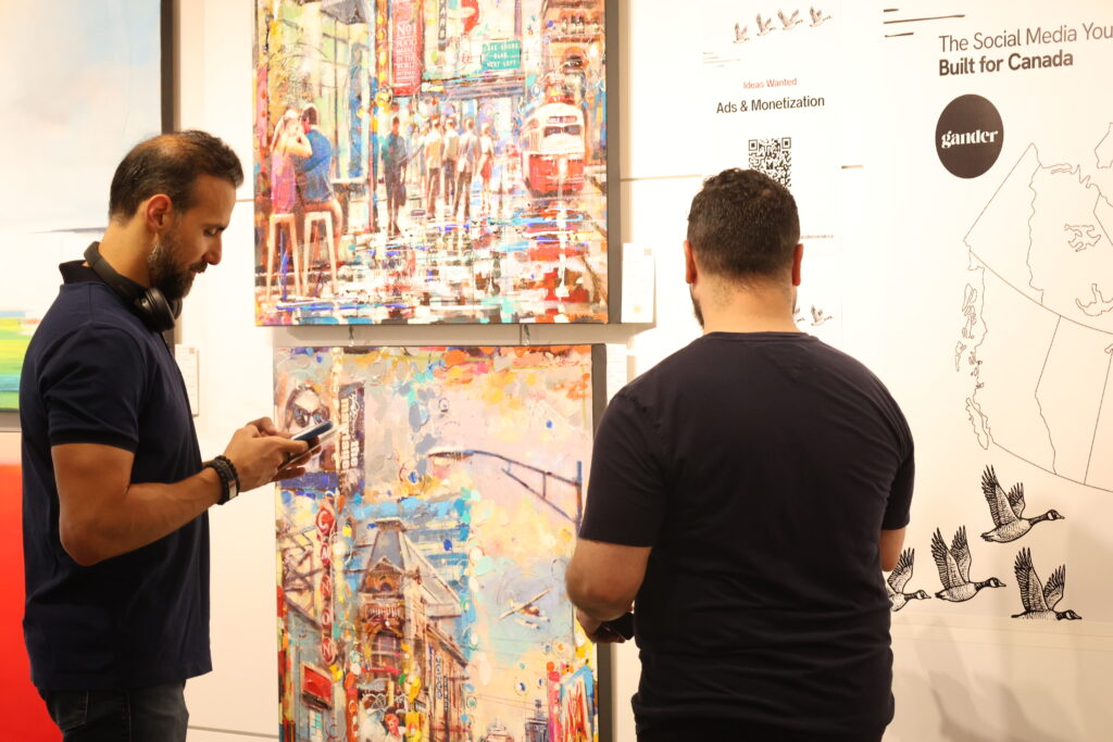 Two men stand in front of colourful cityscape paintings at an art gallery. One looks at his phone while the other faces the artwork. On the wall beside them, Gander branding and a sign reading “Ideas Wanted: Ads & Monetization” are displayed.