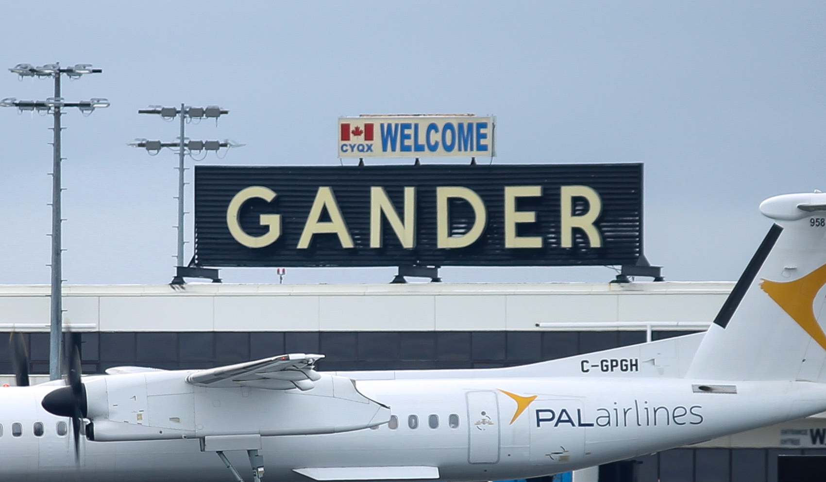 Une enseigne « Welcome Gander » est installée près d'une piste d'aéroport, avec un avion régional au premier plan.