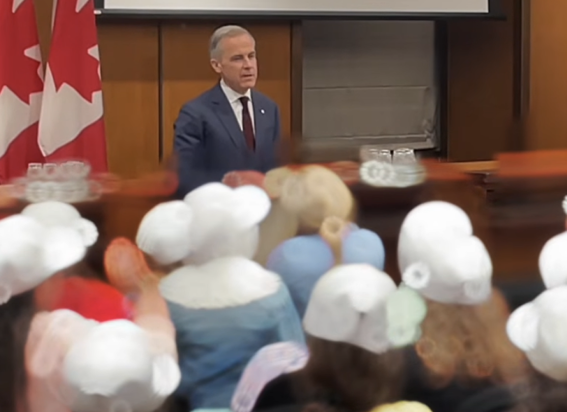 Une personne s'adresse à un auditoire dans une salle de conférence, avec des drapeaux du Canada à l'arrière-plan et des participants assis au premier plan.