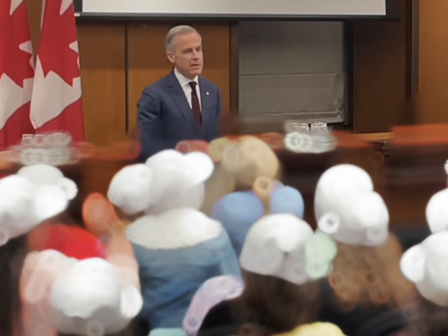 Une personne s'adresse à un auditoire dans une salle de conférence, avec des drapeaux du Canada à l'arrière-plan et des participants assis au premier plan.
